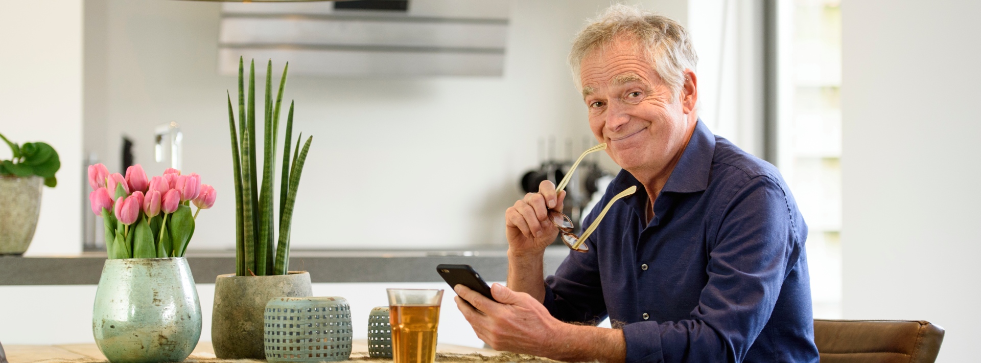 Man met bril in de ene hand en in de andere hand zijn mobiele telefoon zit aan tafel met glas thee