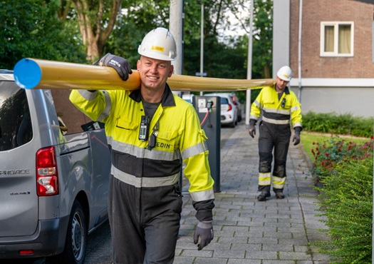 Twee monteurs van Stedin dragen een gele buis lopend door een straat