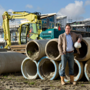 man staat op bouwplaats bij nieuwbouwhuizen met op achtergrond een graafmachine en huizen in aanbouw.