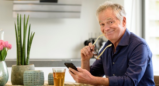 Man aan de keukentafel met mobiel en bril in de hand